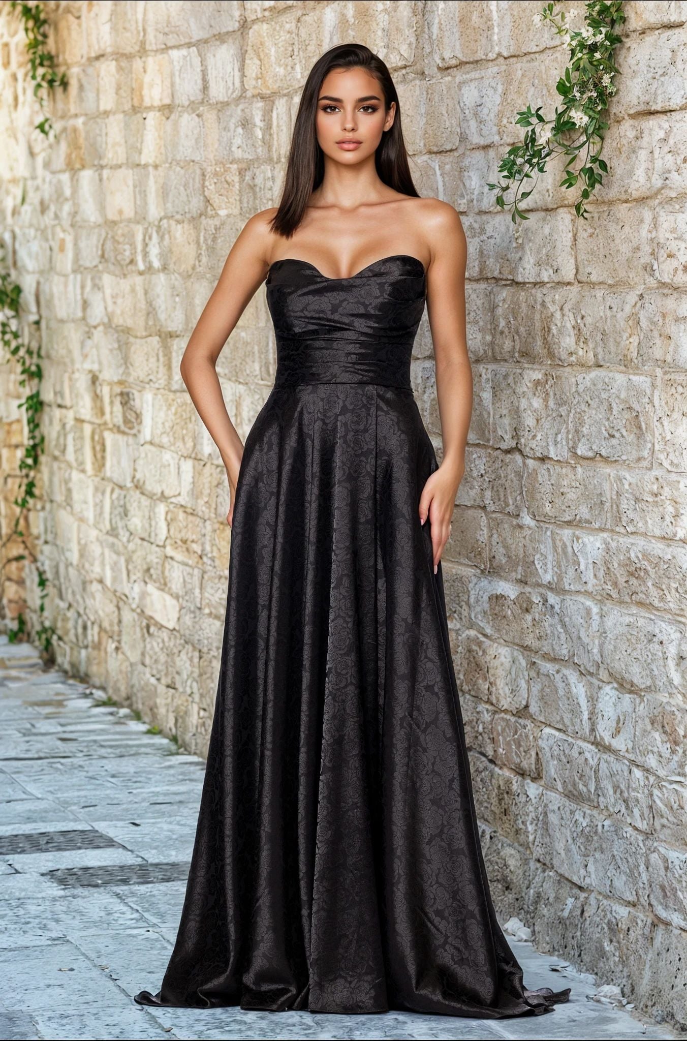 A woman stands against a stone wall, wearing the Strapless Black Prom Gown: Blondie Nites 5301BN—a long black dress with a subtle textured pattern, available in junior sizes for the perfect fit.